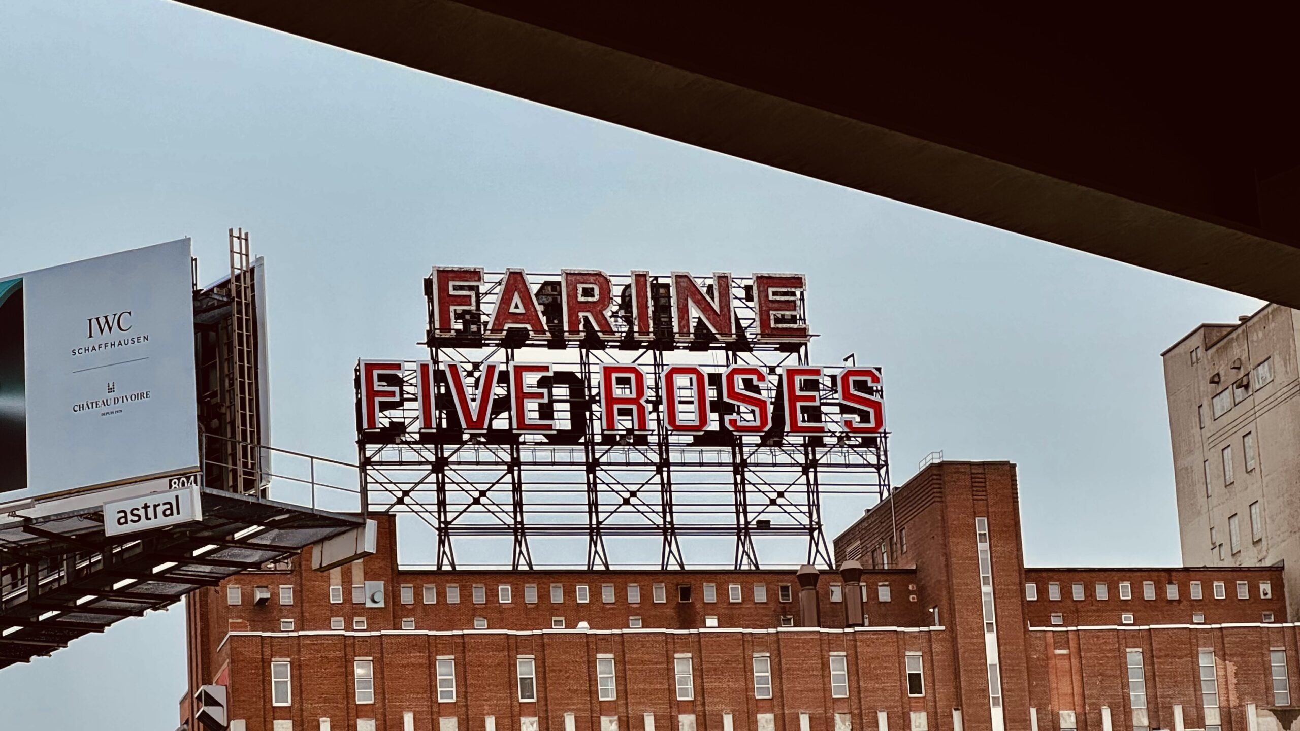 The Farine Five Roses sign photographed from underneath the Bonaventure Expressway, Peel Basin, Tiohtià:ke. The dark underside of the overpass cuts across the top of the frame. The red neon letters rise above a long red brick industrial building against a pale grey sky. An IWC Schaffhausen billboard occupies the left edge.