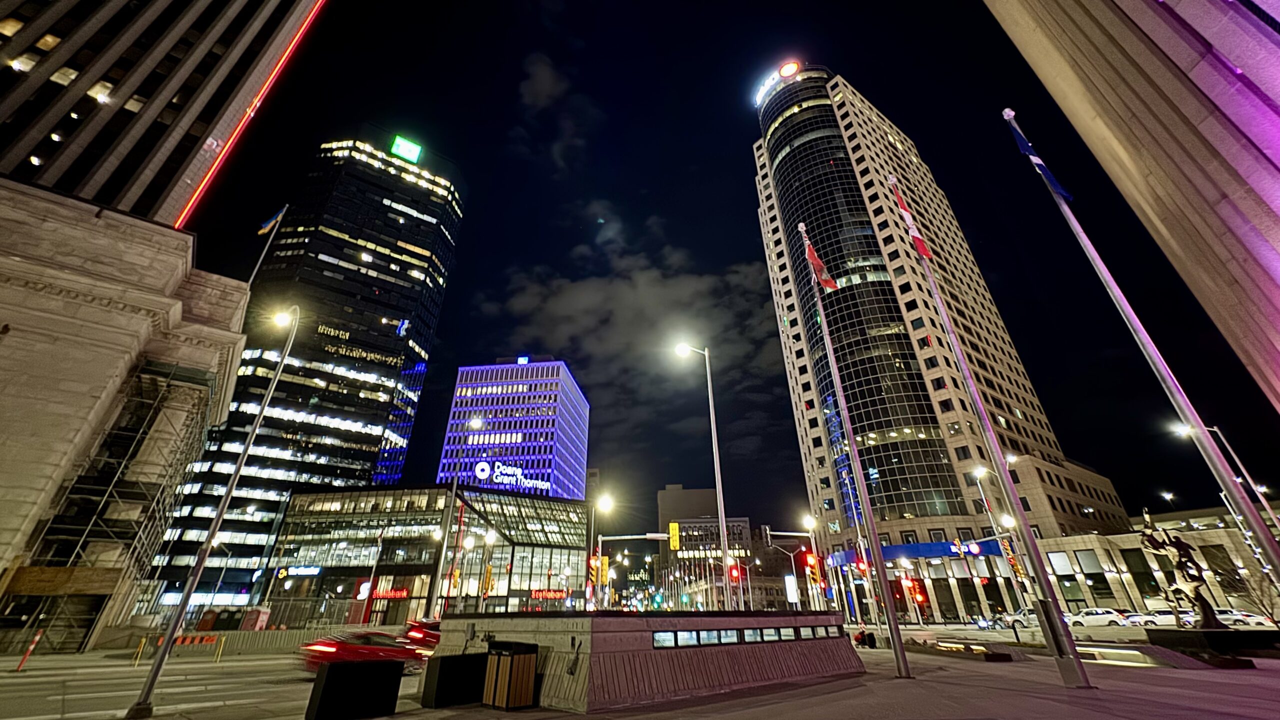 Portage and Main at night, Winnipeg, Treaty One Territory. The Richardson Building curves on the right, the Doane Grant Thornton building glows blue at centre, Canadian flags line the forecourt. The street is largely empty. A Ukrainian flag flies over the Bank of Montreal building on the left of the image. A car's tail lights blur through the intersection and are already gone.