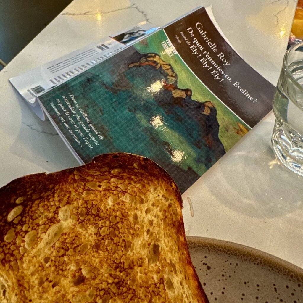 Gabrielle Roy, *De quoi t'ennuies-tu, Éveline? suivi de Ély! Ély! Ély!*, face down and open on a marble counter beside a glass of water and a ceramic mug. A piece of darkly toasted bread in the foreground. The cover painting is a landscape, deep greens and blues, a body of water, land curving at the edge. The back cover text is partially legible in French.
