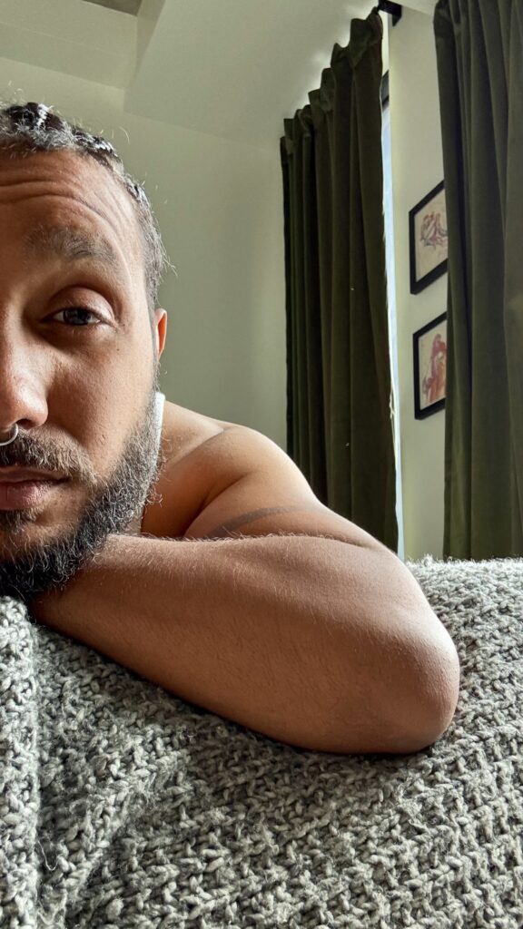 A close, low-angle selfie in soft indoor morning light. Vincent's face fills the left side of the frame, only half visible, eyes looking past the camera rather than into it. Their arm is folded and resting on a textured grey blanket in the foreground, close to the lens. In the background, out of focus, tall olive-green curtains hang beside a window, with two small framed prints on the wall nearby.