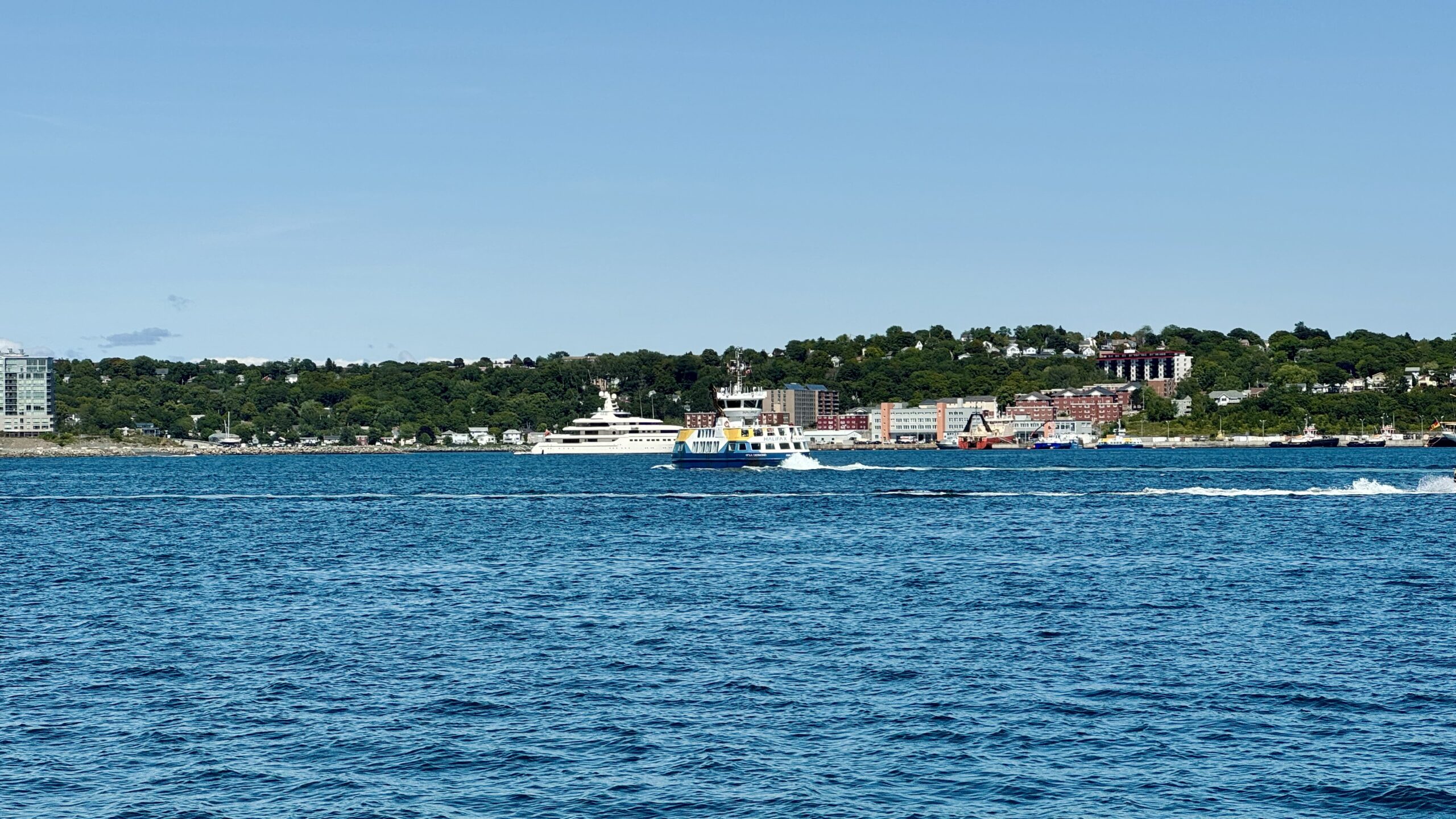 A harbour ferry crosses the water toward the camera, the Dartmouth waterfront and tree-covered hillside behind it under clear sky.